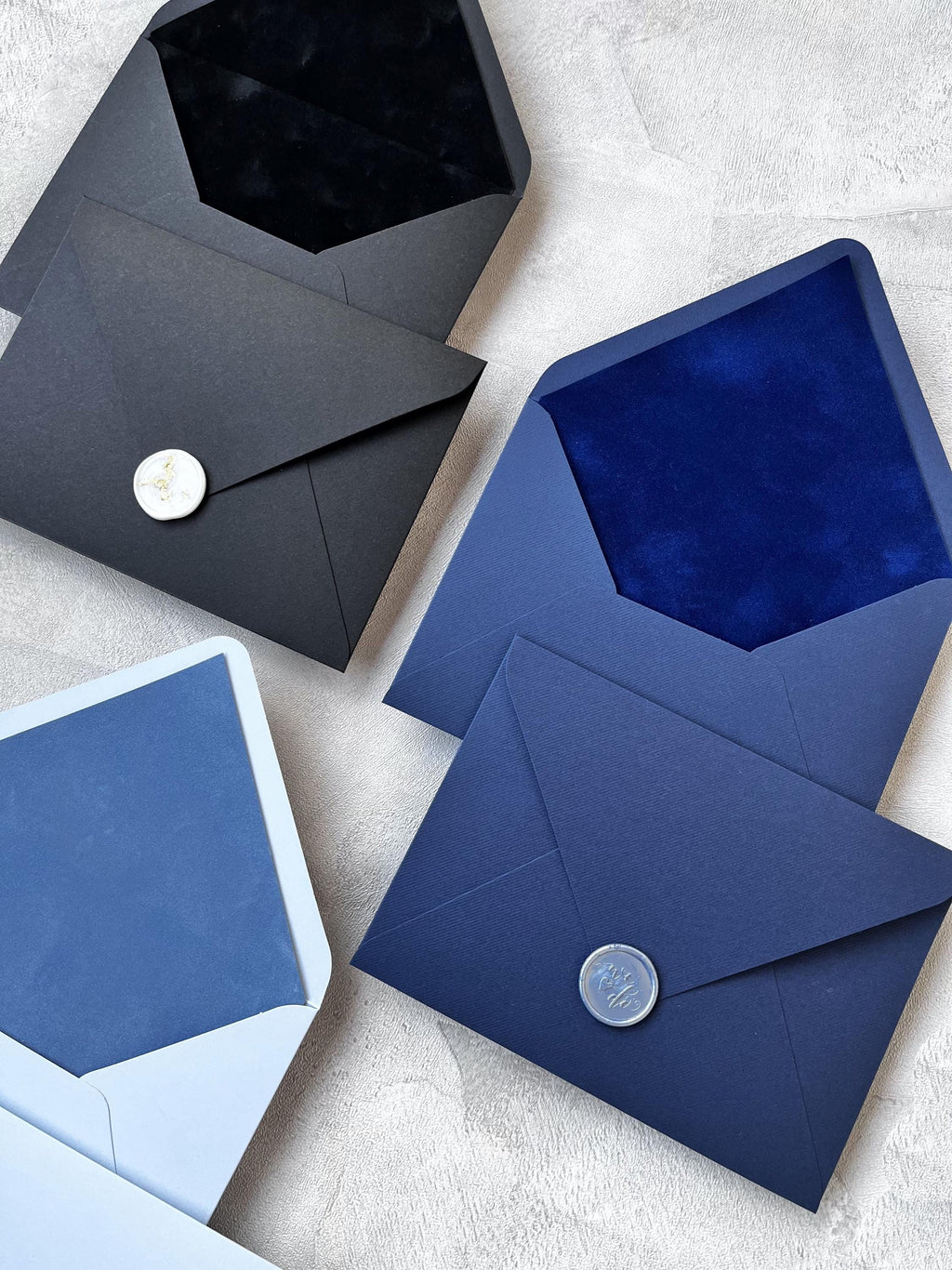 Una collezione di buste in vari colori, tra cui nero, blu navy, azzurro, bianco, crema, oro antico, verde, verde salvia, bordeaux, terracotta e rosa, e rosa cipria. Presentano anche inserti in velluto colorato: nero, blu royal, bianco, verde, pistacchio, crema e bordeaux.
Le buste sono disposte in fila, con la linguetta di ogni busta rivolta verso chi guarda. Tutte le buste sono realizzate in carta spessa e ruvida, i cui colori sono lucidi.