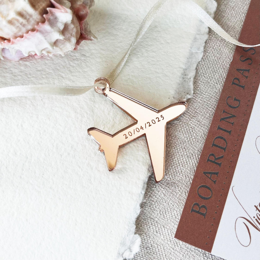 A unique airplane-shaped tag made from mirror gold acrylic with your wedding date elegantly laser engraved. The tag is laced with 3mm pearl satin ribbon for a touch of sophistication. It sits on a printed card with a world map and continents, creating a stunning travel-inspired presentation.