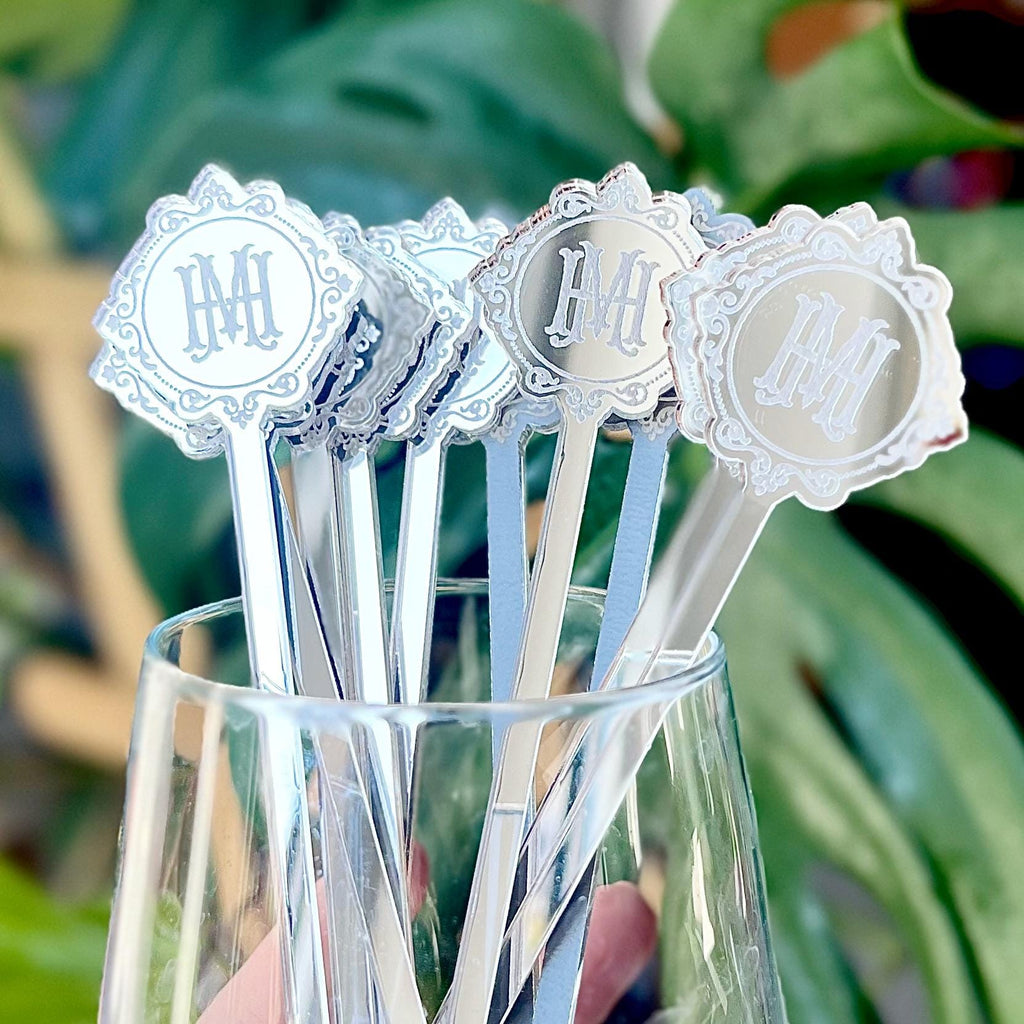 A set of silver, mirror-finish drink stirrers personalized with the bride and groom’s initials in a monogram design. The personalization is done using laser engraving. Around the letters, an elegant decorative pattern is also engraved.