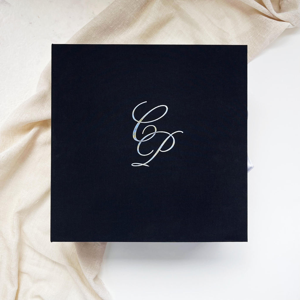 The black linen box is shown open. The left side of the cover shows the wedding venue, embossed with white foil on linen, and the right side has space for 6x9 acrylic or paper invitations and a bottle of wine/champagne. The box has a double-sided white satin ribbon that can be used to tie the box.