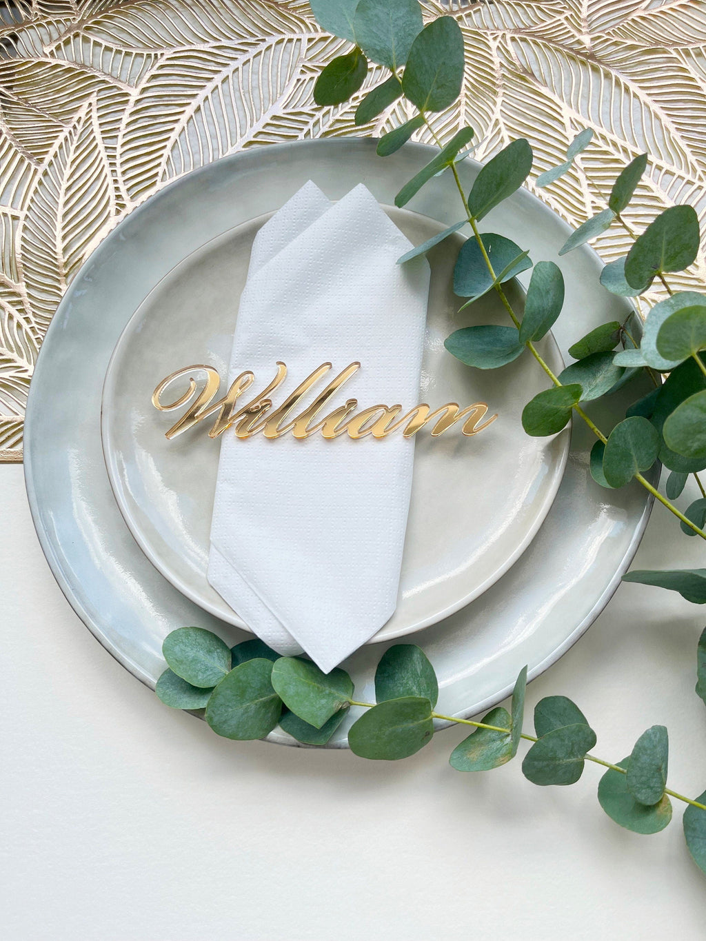 May include: A gold acrylic place card with the name "William" written in a cursive font. The place card is sitting on a white napkin on a grey plate.