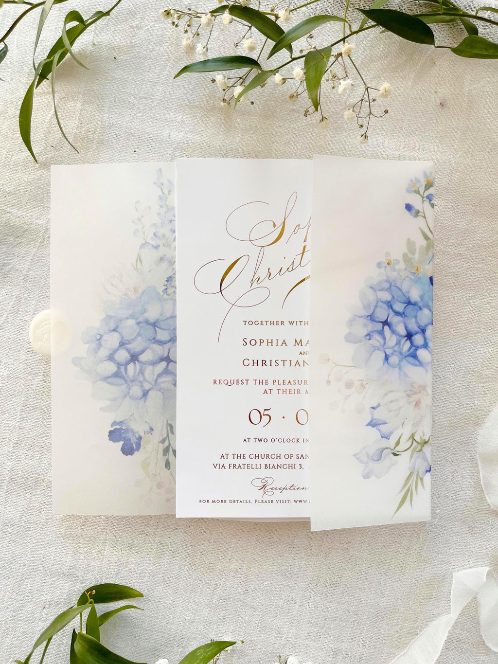May include: White wedding invitations featuring a blue hydrangea floral design. The invitations are sealed with a white wax seal. The design includes blue and white watercolour flowers and greenery. The invitations are on a white linen surface.