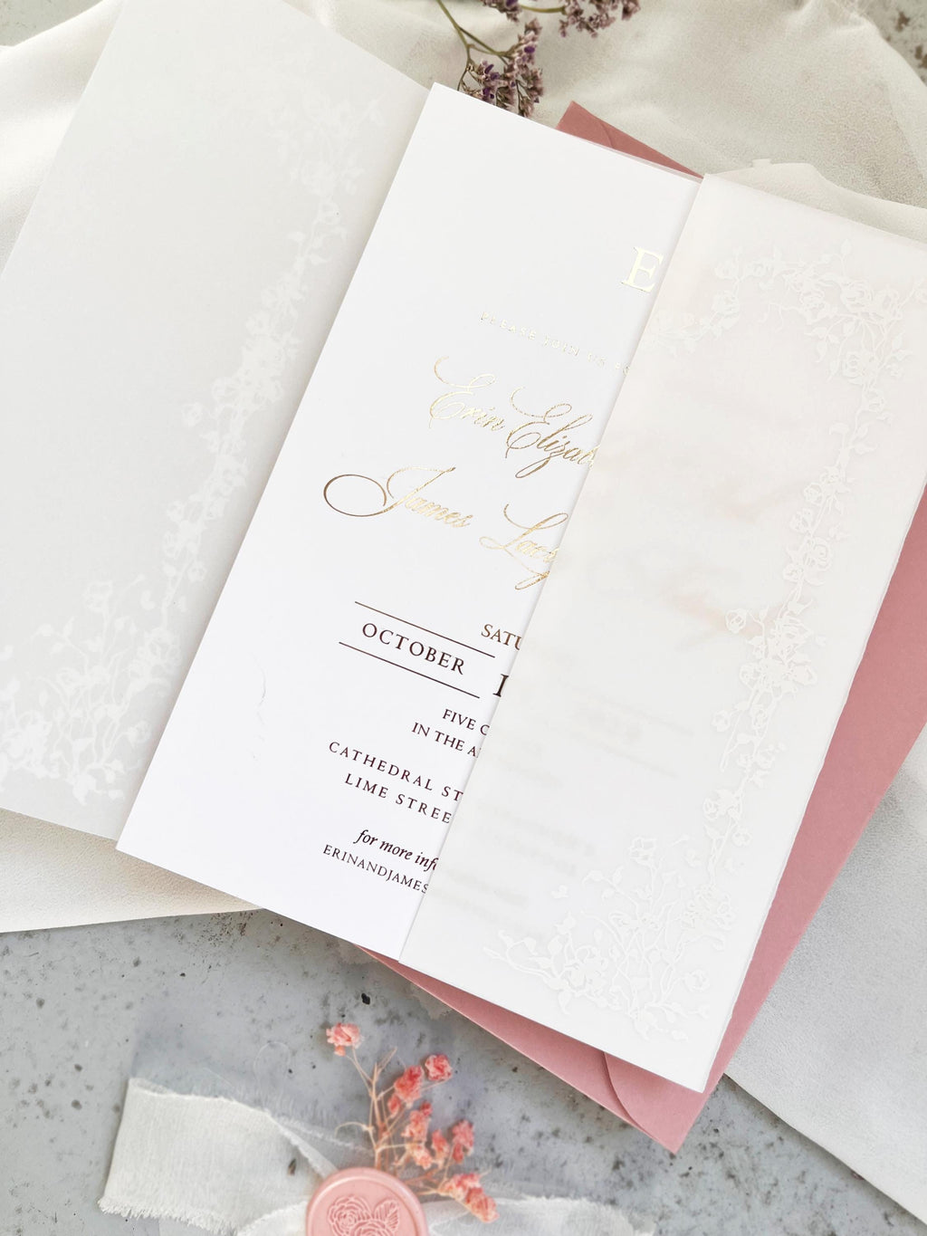 May include: Two white wedding invitations with floral lace designs. One is sealed with a white wax seal. The invitations are on a white fabric background with dried flowers.